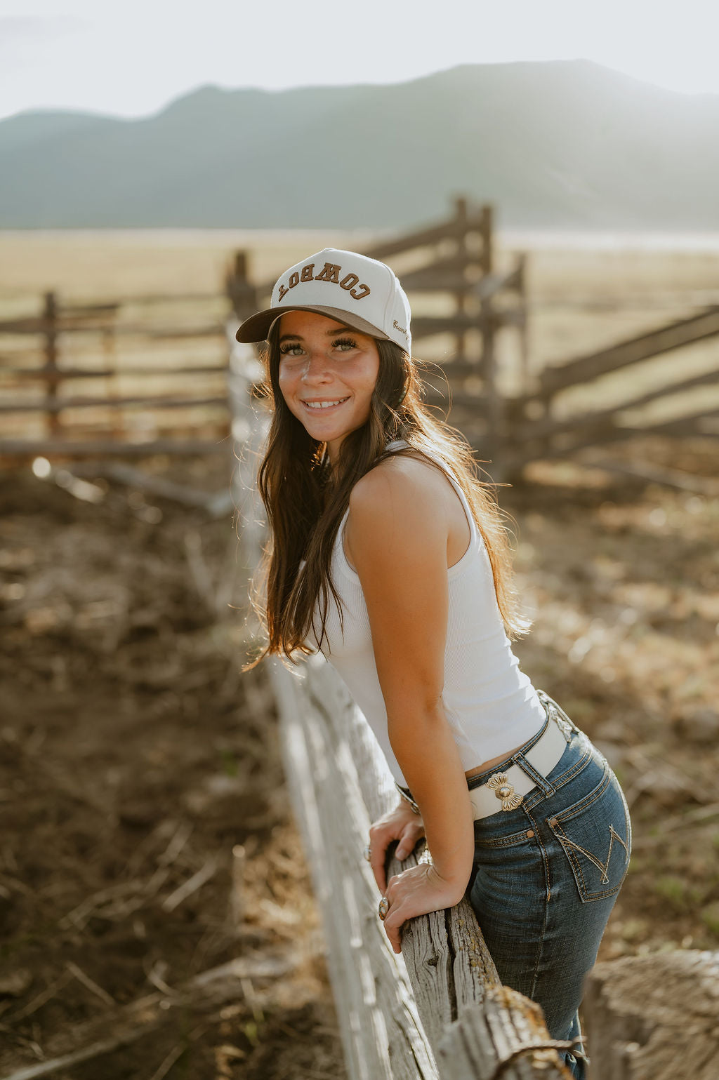 A smiling young woman in a white tank top, blue jeans, and a white baseball cap with 'Cowboy' printed on the front, leaning on a weathered wooden fence in a sunlit outdoor ranch setting with mountains in the background.