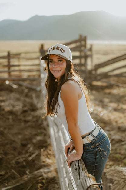A smiling young woman in a white tank top, blue jeans, and a white baseball cap with 'Cowboy' printed on the front, leaning on a weathered wooden fence in a sunlit outdoor ranch setting with mountains in the background.